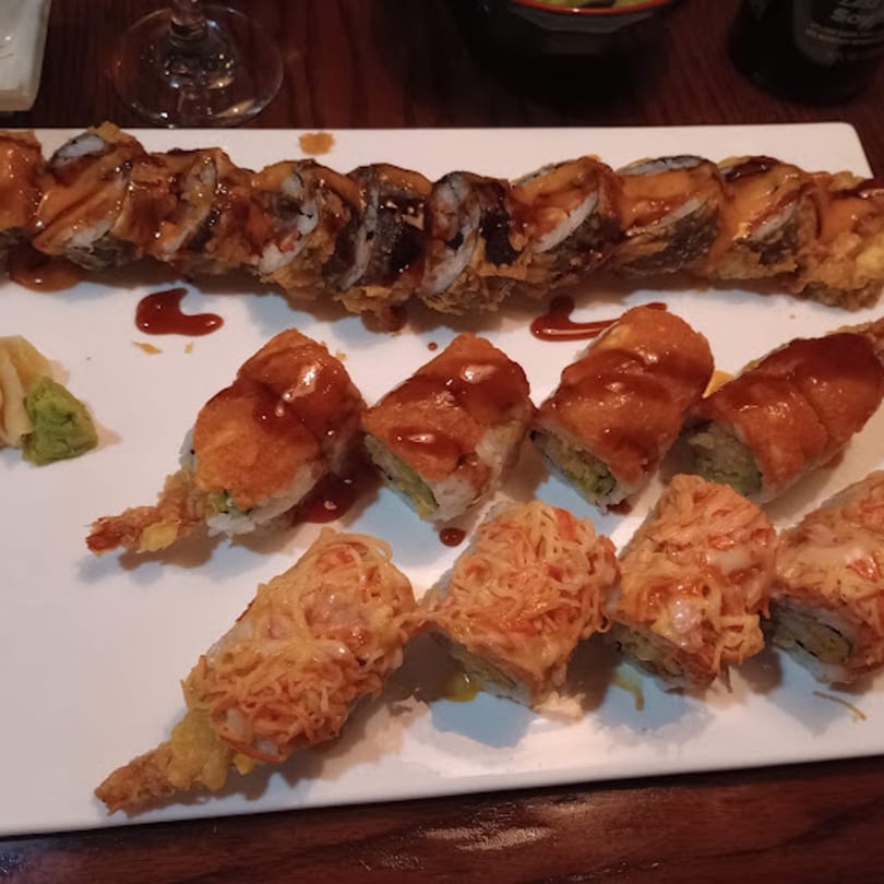Three rows of various sushi rolls, some fried and drizzled with sauce, on a white plate.